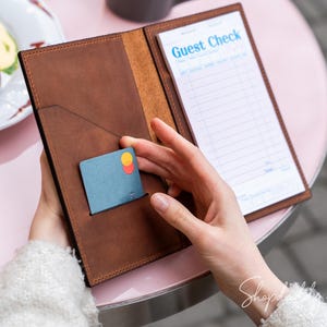 May include: A brown leather guest check holder with a credit card in the front pocket. The holder is open, revealing a notepad with the words "Guest Check" printed on it. The holder is on a pink table.