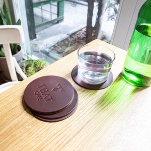Puede incluir: Un conjunto de posavasos de cuero marrón con el texto "THAT CAFE" grabado en relieve en el posavasos superior. Un vaso de agua y una botella de vidrio verde están sobre una mesa de madera clara cerca de una ventana.