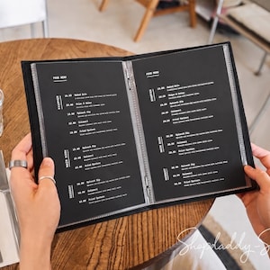 May include: A black restaurant menu folder with clear plastic sleeves containing white text on black pages. The menu lists sections for starters, main dishes, and desserts with prices. The folder is held open, displaying two pages of printed food options.