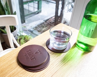 以下が含まれることがあります： 「THAT CAFE」の文字がエンボス加工された茶色の革製コースターのセット。水の入ったグラスと緑色のガラス瓶が、窓の近くにある明るい木製のテーブルの上に置かれています。