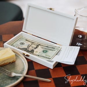 May include: A white rectangular box with a gold clasp, containing a twenty-dollar bill and a receipt, sits on a wooden table. A slice of cheesecake and a fork are on a plate. A glass pitcher with coffee is also on the table.