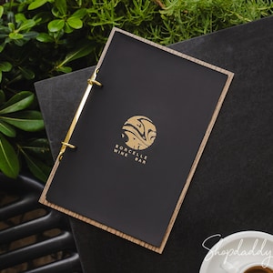 May include: A rectangular menu cover with a matte black front panel and a light wood border. The cover features a gold circular logo with abstract patterns and the text Borcelle Wine Bar. It is held together by two gold metal binder rings.