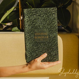 May include: A dark green rectangular journal featuring an embossed leaf-like pattern across the cover. A central rectangular panel displays the gold-colored text CHATEAU BORCELLE 1912. The journal has a textured surface and a vertical orientation.