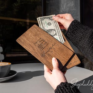 Op de afbeelding: Een rechthoekige donkerbruine houten rekeninghouder met een gladde afwerking. De voorzijde is voorzien van een lasergegraveerd logo en cursieve tekst. De houder staat open en toont een biljet van één dollar in de gleuf aan de binnenzijde.