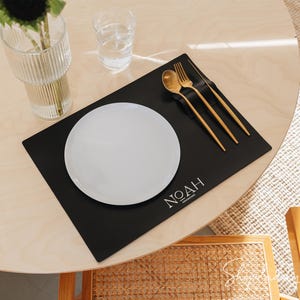 May include: A dining table setting with a black placemat, white plate, and gold cutlery. The placemat has the word "NOAH" printed on it. A clear glass vase with a black flower and a glass of water are also on the table.