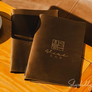 May include: Two brown leather menu covers with gold lettering that reads "Wine Bar" and a wine-themed graphic. The covers are on a wooden table, with a cup of coffee in the background.