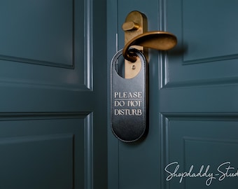 May include: A black leather door hanger with white text that reads PLEASE DO NOT DISTURB, suspended from a brass-colored metal door handle on a dark teal paneled door. The hanger has a smooth, rounded shape with visible stitching along the edges.