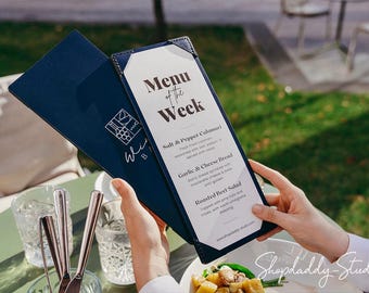 Puede incluir: Una funda de menú de cuero azul oscuro con esquinas cosidas que contiene un inserto blanco titulado Menu of the Week. La cubierta presenta un logotipo gráfico blanco de una copa de vino y uvas. El inserto enumera Salt & Pepper Calamari, Garlic & Cheese Bread y Roasted Beet Salad.