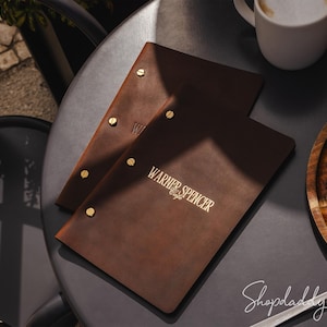 Puede incluir: Dos cubiertas de menú de cuero marrón con detalles dorados y el texto "WARNER & SPENCER Cafe" están sobre una mesa oscura. Una taza de café y una porción de pastel de chocolate también están sobre la mesa.
