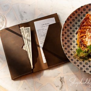 Op de afbeelding: Een donkerbruine leren rekeningmap met twee binnenvakken, waarvan er één bankbiljetten bevat en de andere een geprinte kassabon van Cafe Marko, geplaatst naast een bord met een patroon met eten en een wijnglas.