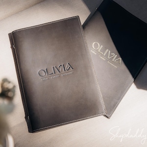 Op de afbeelding: Twee grijze lederen menukaften met het woord "Olivia" in wit reliëf. De tekst "Italiaans restaurant" is ook in reliëf op een van de kaften.