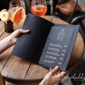 May include: A black menu folder with gold ring bindings, featuring white text and a line-art illustration of wine bottles and glasses. The menu lists items including Dolce Latte Gnocchi and Prawn Malabar Curry with corresponding prices.