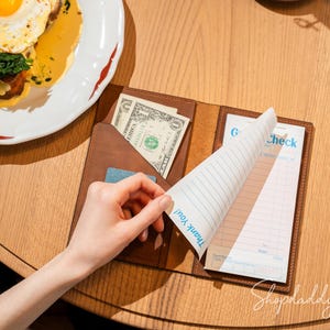 May include: A brown leather server check presenter with a pocket containing a one-dollar bill and a guest check pad. The pad features blue text that reads Guest Check and Thank You on the bottom corner of the top sheet, with lined paper.