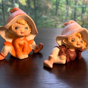 May include: Two ceramic figurines of elves wearing pink hats. The elf on the left is sitting with its legs crossed and wearing an orange outfit. The elf on the right is lying down with its head resting on its hand and wearing a brown outfit.