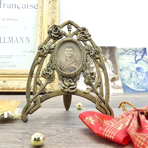 May include: An antique brass-colored photo frame with a vintage portrait. The frame features a floral rose design and an arched top. The frame is supported by a small stand and is displayed on a wooden surface.