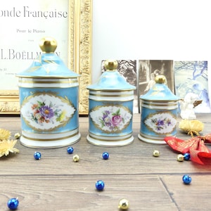 Op de afbeelding: Drie bijpassende blauw-witte porseleinen potten met deksels. Elke pot heeft een bloemenontwerp in een goudomrand ovaal. De deksels zijn bekroond met gouden knoppen. Decoratieve items voor in huis.