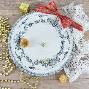 Può includere: Un piatto in ceramica bianca con un motivo floreale blu e un nastro rosso con note musicali. Il piatto è decorato con perline dorate e fiori secchi, ed è circondato da perline dorate e pizzo bianco.