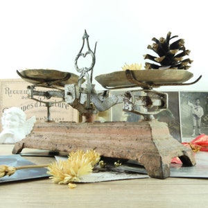 Peut inclure: Balance ancienne en métal blanc et brun avec deux plateaux ronds en laiton. La balance est posée sur une surface en bois avec une pomme de pin et des fleurs séchées.