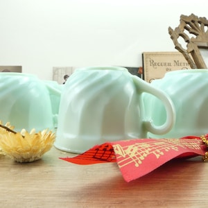 May include: Several light green teacups with a swirled design are arranged on a wooden surface. The teacups have a handle and a rounded base. A red ribbon with gold musical notes and a dried yellow flower are in the foreground.