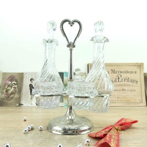 Könnte beinhalten: Ein Servierstück aus klarem Glas und Silber mit zwei Dekantern und zwei kleinen Schalen. Das Stück hat einen herzförmigen Griff und einen dekorativen Sockel. Die Dekanter haben ein gedrehtes Design und die Schalen haben einen gemusterten Rand. Das Stück steht auf einer Holzoberfläche.