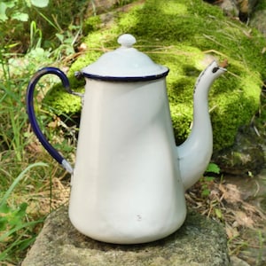 Puede incluir: Cafetera de esmalte blanco con borde y asa azules. La cafetera tiene un cuerpo redondeado y un pico en el lateral.