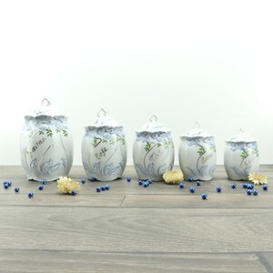 Peut inclure: Cinq pots en céramique blanche avec couvercles, ornés de motifs floraux bleus et d'étiquettes manuscrites. Les pots sont disposés sur une surface en bois, avec des perles bleues et des fleurs séchées éparpillées.