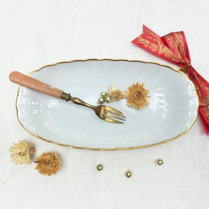 Peut inclure: Un plat de service blanc de forme ovale avec un bord doré, contenant une fourchette vintage avec un manche marron clair. Des fleurs séchées et des perles dorées sont disposées sur le plat. Un ruban rouge avec du texte doré est attaché sur le côté.