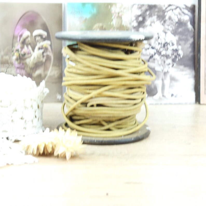 May include: A spool of gold-colored cord or wire on a black plastic spool. The spool is sitting on a wooden surface with some white lace and a few decorative items.