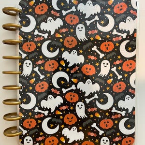 May include: A Halloween-themed planner with a black background and a repeating pattern of orange pumpkins, white ghosts, crescent moons, candy corn, and bone shapes. The planner has gold-colored disc binding.