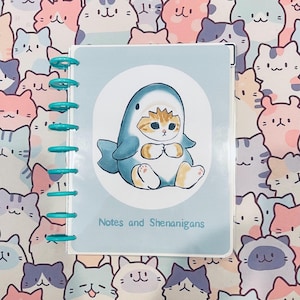 May include: A blue and white shark costume with a cute orange and white cat wearing it. The costume has a fin on the back and a tail. The cat is holding its paws together in front of its chest. The image is on a light blue background with the text "Notes and Shenanigans".