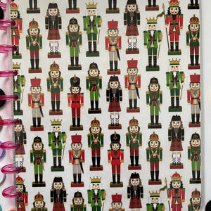 May include: A white background with a repeating pattern of nutcracker figurines. The nutcrackers are all different, with some wearing red and green uniforms, some wearing black and red uniforms, and some wearing blue and gold uniforms. They all have gold crowns and hold swords or staffs.