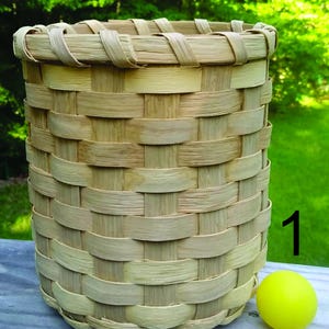 May include: A handcrafted, cylindrical basket woven from light-colored, natural wood strips. The basket has a rounded rim and a sturdy, woven construction. A small, yellow ball sits next to the basket. The number 1 is visible on the right side of the basket.