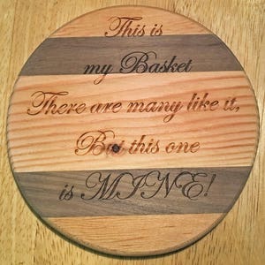 May include: A round wooden cutting board with a striped design in brown and light wood. The board is engraved with the text "This is my Basket There are many like it, But this one is MINE!"