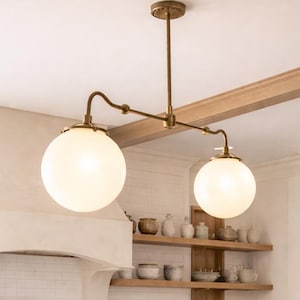 May include: A bright, airy kitchen with a marble-topped island and woven-back bar stools. Overhead, a brass light fixture with two globe lights illuminates the space. A wooden cutting board with bread and lemons sits on the island, and a vase of flowers adds a fresh touch.