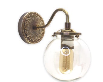 May include: A brass wall sconce with a clear glass globe shade and a decorative backplate. The sconce has a curved arm and a single light bulb.