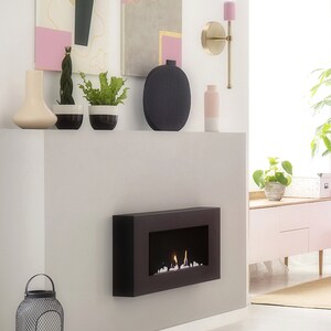 May include: A modern interior scene featuring a black fireplace with flames, a decorative wall sconce, and abstract art. The room is decorated with vases, plants, and a metal lantern. The color palette includes black, pink, gold, and white.