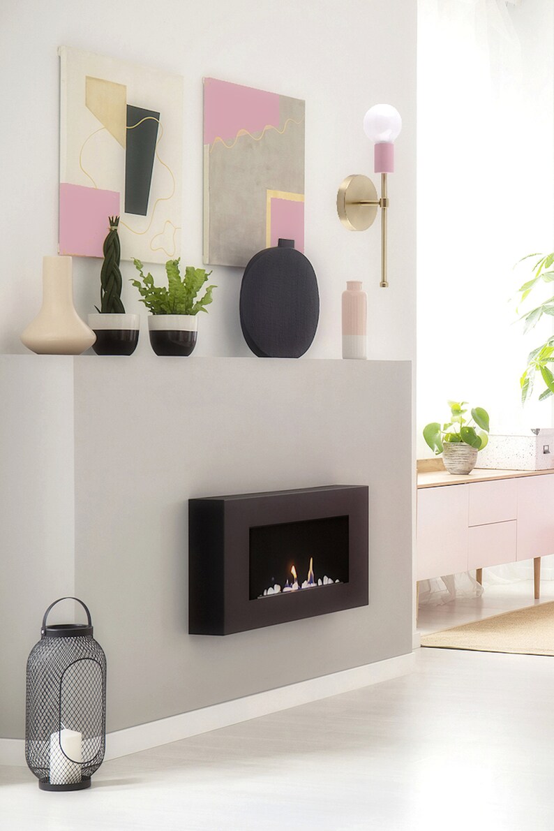 May include: A modern interior scene featuring a black fireplace with flames, a decorative wall sconce, and abstract art. The room is decorated with vases, plants, and a metal lantern. The color palette includes black, pink, gold, and white.