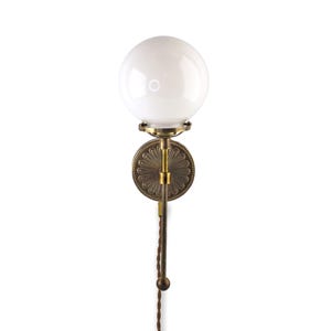 May include: A brass wall sconce with a white globe shade and a decorative floral design on the back plate. The sconce has a long, thin arm with a cord hanging down.