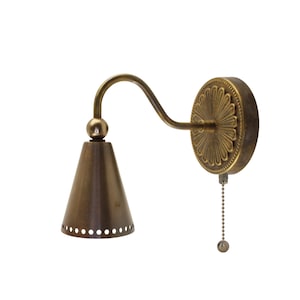 May include: Antique brass wall sconce with a cone-shaped shade and decorative floral backplate. The sconce features a curved arm and a pull chain for easy operation. The shade has small circular perforations.