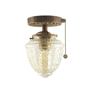 May include: A brass ceiling light fitting with a clear glass shade featuring a textured pattern. The light has a pull chain.