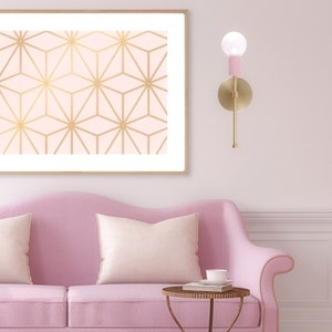 May include: A pink sofa with two cream-colored pillows and a small, round, antique-style table. A framed print with a gold geometric pattern hangs above the sofa. A gold sconce with a pink accent and a light bulb is mounted on the wall.