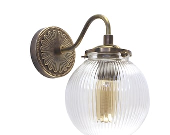 May include: A brass wall sconce with a clear ribbed glass globe shade. The sconce has a decorative floral design on the back plate.