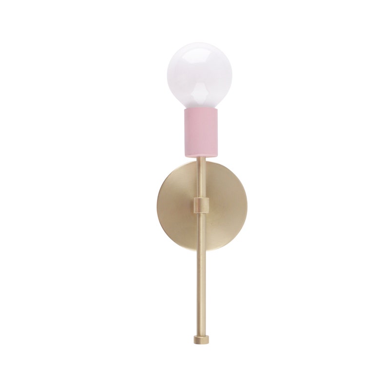 May include: A modern wall sconce with a gold-toned metal finish. It features a round backplate, a slender stem, and a pink cylindrical accent holding a white bulb. The design is minimalist and elegant.