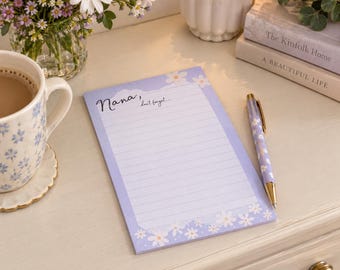 May include: A rectangular notepad with a light purple background featuring white daisy patterns at the top and bottom corners. The notepad has horizontal lines and the printed text "Nana, don't forget..." at the top. A matching purple daisy pen rests beside it.