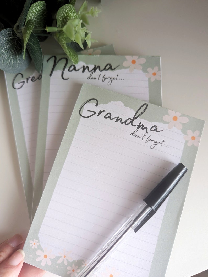 May include: Three green and white notepad sets with a floral design. The notepads have the words "Grandma", "Nanna", and "Great" written on them. Each notepad has the text "don't forget..." written on it. A black pen is lying on top of the "Grandma" notepad.