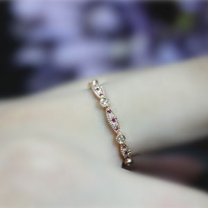 May include: A thin gold band ring featuring a repeating pattern of marquise-shaped and circular settings. The marquise sections contain small red stones, while the circular settings are accented with clear white stones, all set in yellow gold.