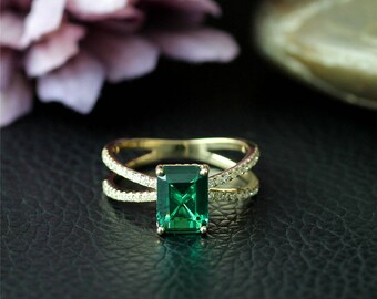 May include: A gold ring with a large emerald green square-shaped gemstone surrounded by smaller clear gemstones. The ring has a split band design.
