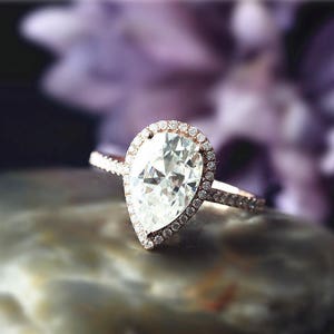 Może przedstawiać: Pierścionek zaręczynowy z diamentem w kształcie gruszki z oprawą halo. Pierścionek jest wykonany ze złota różowego i ma jeden duży diament w środku.