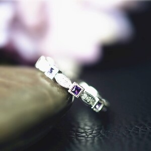 Puede incluir: Un anillo de tono plateado con un patrón repetitivo de piedras preciosas moradas de corte cuadrado que se alternan con pequeños segmentos metálicos ovalados y texturizados. La banda tiene un detalle de borde milgrain alrededor de cada engaste.