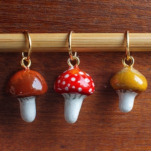 Puede incluir: Tres pendientes con forma de seta con aros dorados. Uno es marrón, otro rojo con lunares blancos y el otro amarillo. Cada uno tiene un tallo blanco. Los pendientes se muestran en una clavija de madera sobre un fondo de madera.